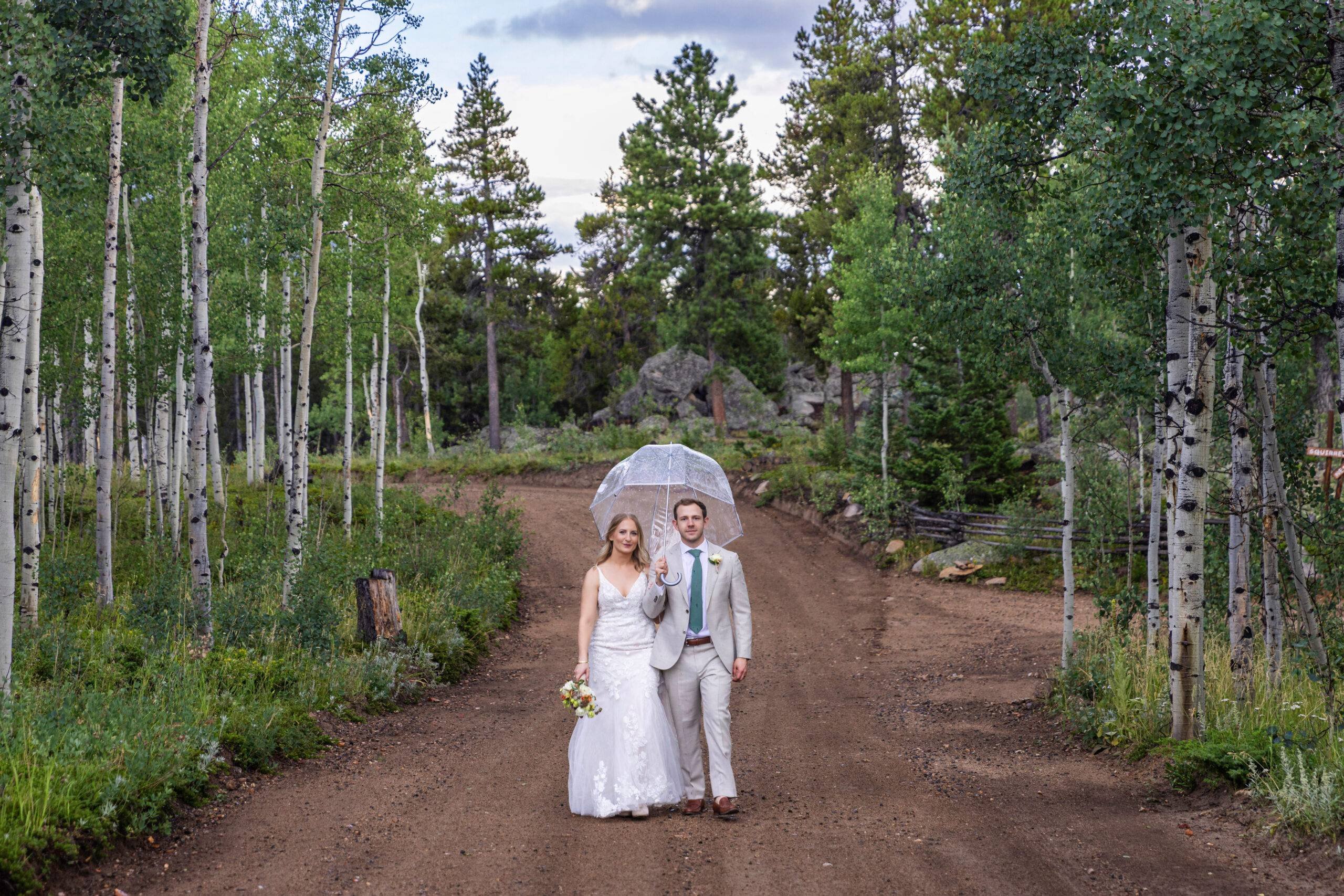A bride and groom stand on a dirt road in Colorado with trees surrounding them.