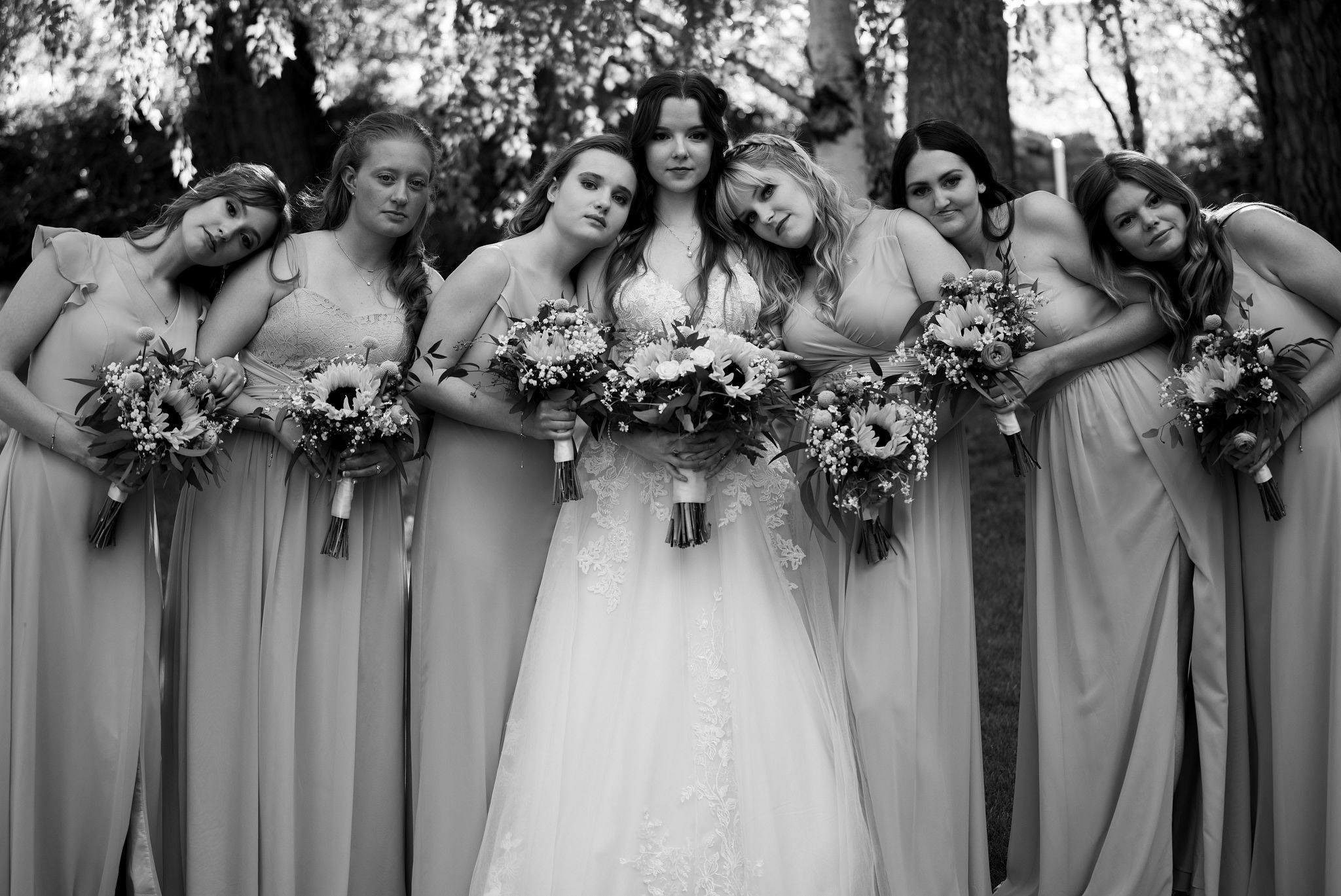 black and white photo of a bride surrounded by six bridesmaids. The group stands closely together outdoors, with the bridesmaids leaning affectionately toward the bride. Each woman holds a bouquet of flowers featuring sunflowers and greenery. The bride wears a lace wedding gown, while the bridesmaids wear matching long dresses. Trees and dappled sunlight form the background.